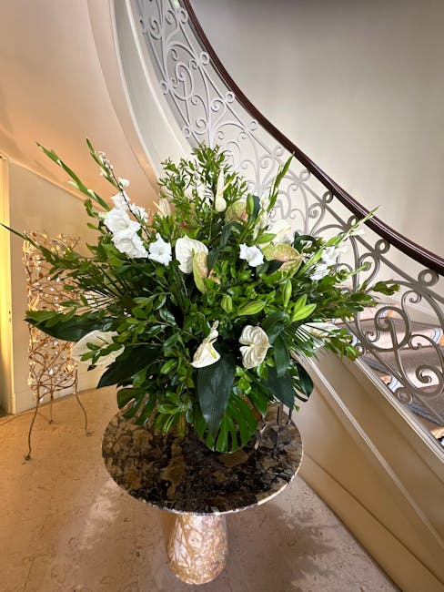 A large, elegant floral arrangement housed in a black vase sits on a polished marble pedestal table inside a building with ornate white railings and soft, ambient lighting. The bouquet features white 