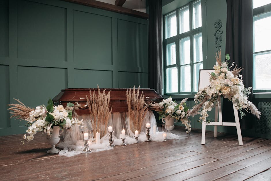 A floral arrangement featuring white roses, lilies, and orchids complemented by dried pampas grass and greenery, arranged in two white ceramic vases. The flowers are meticulously placed on a wooden fl