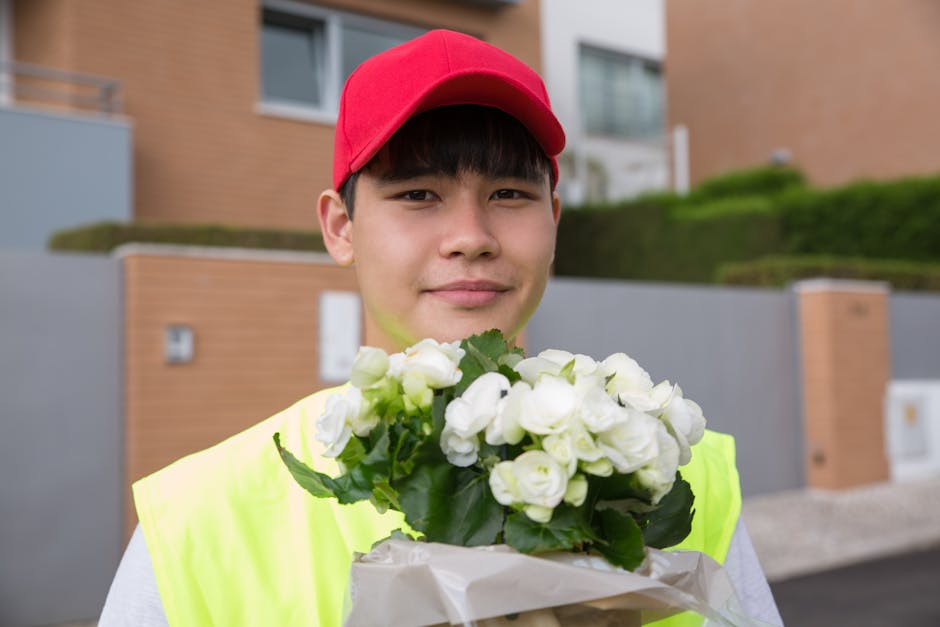 A young male flower delivery courier with light skin, wearing a red cap and neon yellow high-visibility vest, stands outdoors in front of modern brick residential buildings with greenery and bushes. H