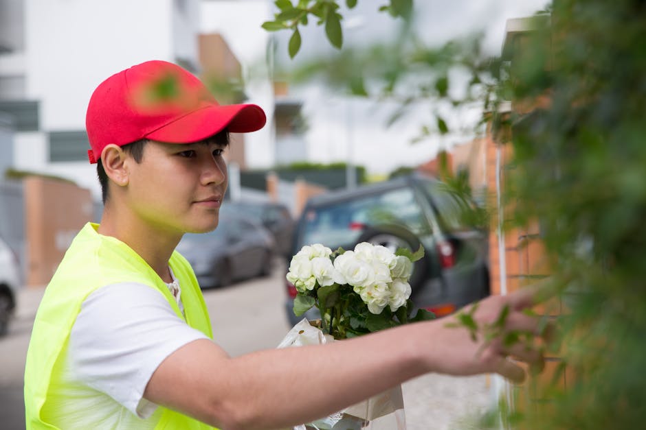 A young male florist wearing a red cap and a neon yellow vest delivers a floral gift in Fulham. He holds a bouquet of fresh white hydrangeas wrapped in white paper, carefully reaching to hang the flow