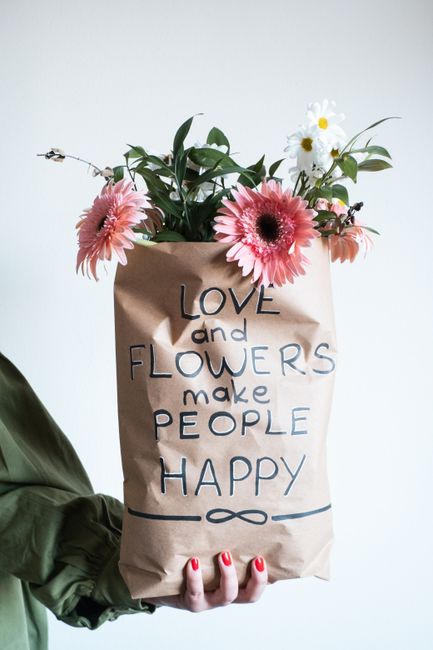 A person wearing a dark green jacket is holding a brown paper bag filled with an arrangement of fresh flowers, including pink gerbera daisies, white daisies with yellow centers, and green foliage. The